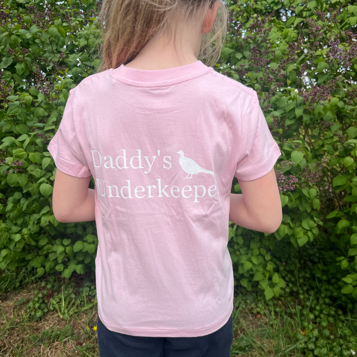 Child wearing a pink t-shirt with 'Daddy's Underkeeper' text, standing outdoors.